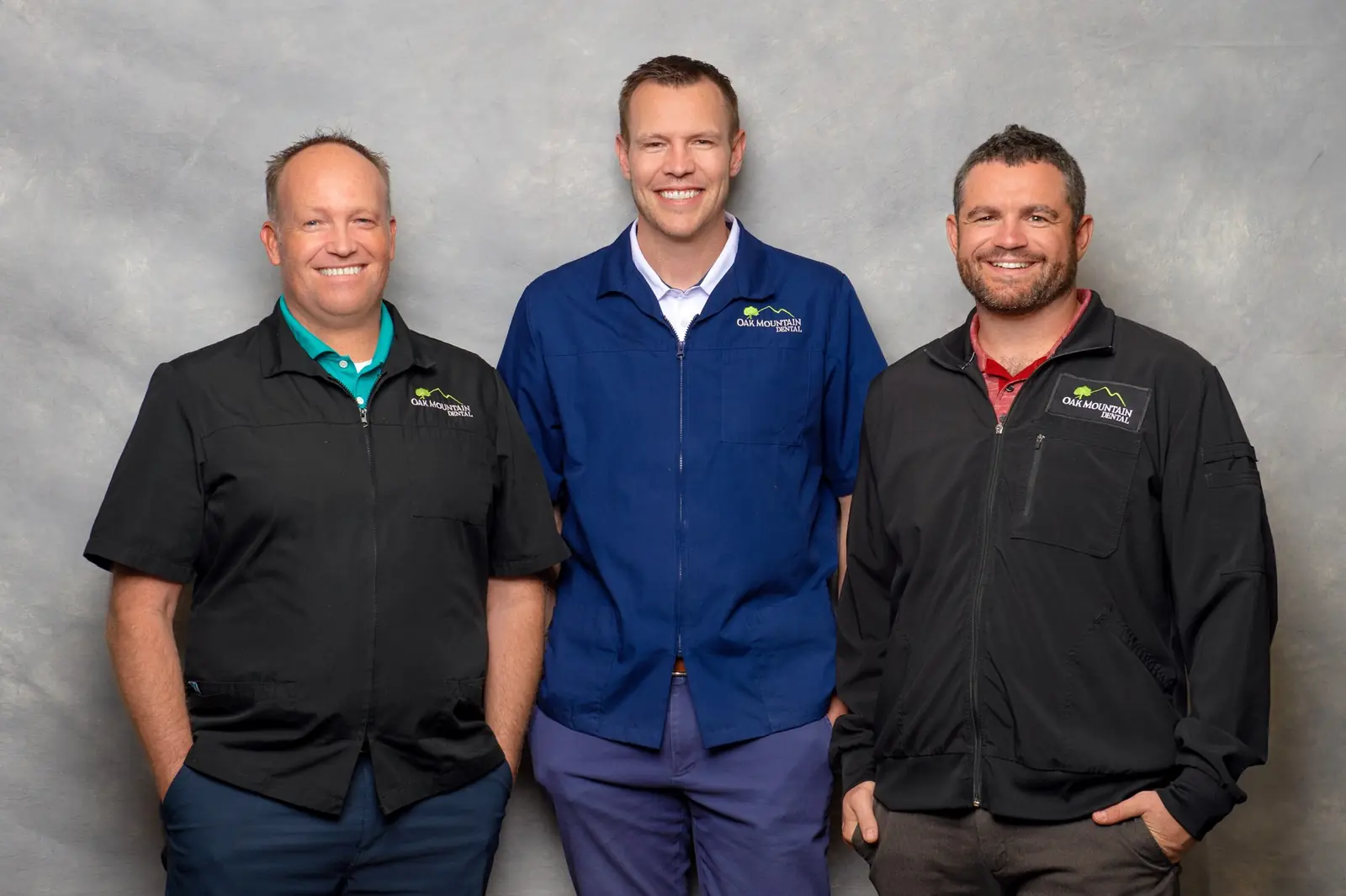 Three smiling dental professionals wearing Oak Mountain Dental uniforms in front of a gray backdrop.