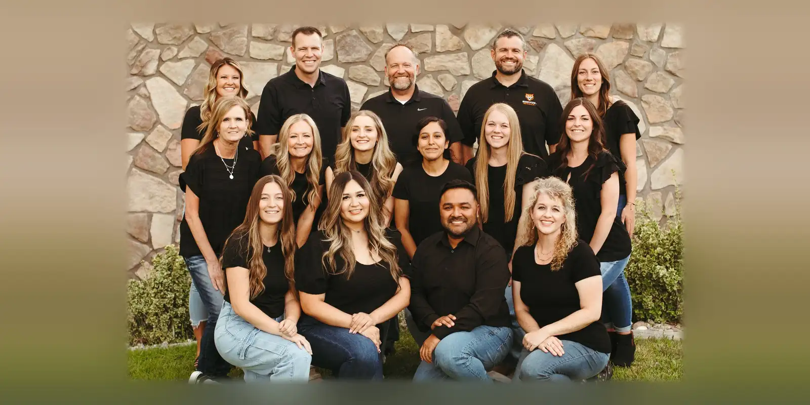 A group of dental team members smiling outdoors in front of a stone wall, wearing black tops and jeans.